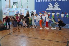 Na tle dekoracji grupa dzieci przebrana za postaci z wiersza. Dzieci stoją jedno za drugim i trzymają się w pasie. Próbują ,,wyciągnąć” tytułową rzepkę/rekwizyt wykonany z bibuły leży na podłodze przed pierwszym dzieckiem.