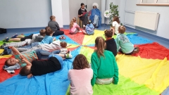 Grupa uczniów siedzi na kolorowej chuście na podłodze, a dwoje uczniów z klasy ósmej czyta im bajkę.