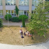 Ujęcie z lotu ptaka na grupę uczniów czytających przed budynkiem szkoły.