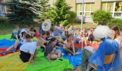 Grupa uczniów siedzi na kolorowej chuście na trawie, a dwoje uczniów z klasy ósmej czyta im bajkę.