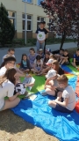 Grupa uczniów siedzi na trawie na kolorowej chuście, a dwoje uczniów z klasy ósmej czyta im bajkę.