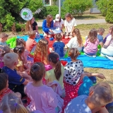 Kolorowa grupa dzieci siedzi na trawie i słucha bajki czytanej przez cztery starsze koleżanki.