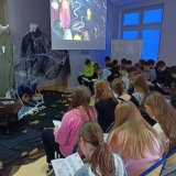 Grupa uczniów czyta II część „Dziadów” Adama Mickiewicza w scenerii biblioteki szkolnej zaaranżowanej specjalnie na tę okoliczność.