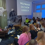 Grupa uczniów czyta II część „Dziadów” Adama Mickiewicza w scenerii biblioteki szkolnej zaaranżowanej specjalnie na tę okoliczność.