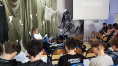 Grupa uczniów czyta II część „Dziadów” Adama Mickiewicza w scenerii biblioteki szkolnej zaaranżowanej specjalnie na tę okoliczność.