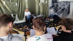 Grupa uczniów czyta II część „Dziadów” Adama Mickiewicza w scenerii biblioteki szkolnej zaaranżowanej specjalnie na tę okoliczność.