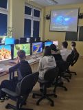 Dzieci w sali z komputerami, uczestniczące w zajęciach z bezpieczeństwa w internecie, oglądające prezentację o tematyce "Bezpieczny Internet"