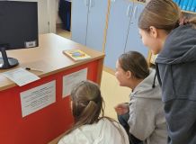 Trójka nastolatków uważnie czyta jedno z zadań w szkolnej bibliotece, obok biurko z książkami i dokumentami szkolnymi.