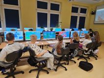 Klasa z dziećmi grającymi w edukacyjną grę Interelandia na komputerach stacjonarnych, dobrze oświetlone środowisko szkolne z dużymi oknami i wzorami na ścianach.