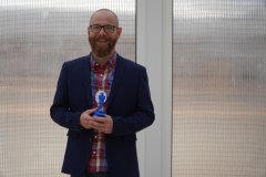 Najpopularniejszy Nauczyciel, Pan Łukasz Grocki- w ręku trzyma wydrukowaną na drukarce 3D niebieską statuetkę, a na szyi zawieszony ma biały medal.