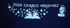 Dekoracja – na czarnej kurtynie biały napis Dzień Edukacji Narodowej, otwarta księga, z której wylatują motyle, z prawej strony sowa w birecie.