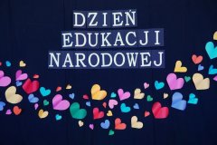 Na ciemnym tle widnieje biały napis „Dzień Edukacji Narodowej”. Poniżej znajduje się dekoracja wykonana z kolorowych serc.