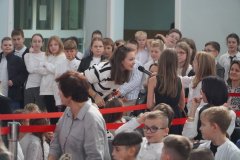 Pani Nauczycielka w białym swetrze w czarne paski odpowiada, do mikrofonu, na pytanie. W tle znajdują się  Uczniowie w biało-czarnych strojach galowych.
