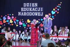 Dyrektor Szkoły- p. Marlena Błachowicz, ubrana w czerwoną sukienkę,  oraz Przewodniczący Rady Rodziców- p. Przemysław Zieliński , ubrany w szary garnitur,  przemawiają do mikrofonu , stojąc na tle dekoracji wykonanej z kolorowych serc i białego napisu „Dzień Edukacji Narodowej”.
