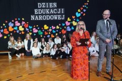 Dyrektor Szkoły- p. Marlena Błachowicz, oraz Przewodniczący Rady Rodziców- p. Przemysław Zieliński, przemawiają z wykorzystaniem mikrofonów. W tle znajduje się chór, a za nimi dekoracja wykonana z kolorowych serc dopełniona białym napisem „Dzień Edukacji Narodowej”.