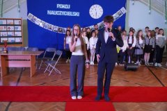 Na tle granatowej dekoracji z napisem pokój nauczycielski, dwójka narratorów stojących na czerwonym dywanie – Justyna i Adam rozpoczynają część artystyczną akademii.