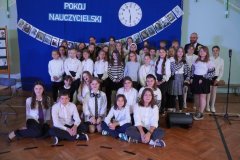 Na tle granatowej dekoracji z napisem pokój nauczycielski stoją uczniowie śpiewający w chórze TuTu oraz zespół muzyczny.