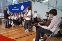 Na pierwszym planie zespół muzyczny grający na gitarach, perkusji i keyboardzie, w tle śpiewający chór TuTu przy batucie pana Łukasza Grockiego.