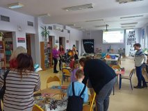we wnętrzu biblioteki widać grupę rodziców i dzieci, bibliotekarze prezentują dzieciom i rodzicom nietypowe książki: pop-up, z rozszerzoną rzeczywistością, itp.