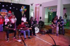 Dzień otwarty w szkole: występ zespołu rockowego na sali gimnastycznej. Uczniowie grają na gitarach elektrycznych, basowych i perkusji. Obok nich stoi grupa wokalna, a w tle widać kolorowe balony, dekoracje z zimowym miasteczkiem oraz drabinki gimnastyczne.