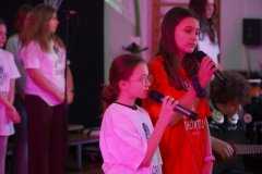 Dzień otwarty w szkole: zbliżenie na dwie dziewczynki śpiewające do mikrofonów podczas występu chóru. Jedna z nich ma na sobie białą, a druga czerwoną koszulkę z logo „Chór Tutu”. W tle widać pozostałych członków zespołu i fragmenty instrumentów.