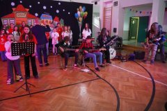 Dzień otwarty w szkole: szerokie ujęcie występu artystycznego w sali gimnastycznej. Na scenie występuje zespół muzyczny z gitarzystami oraz chór szkolny pod okiem nauczyciela muzyki. W tle widać kolorowe dekoracje, pęk balonów i drabinki gimnastyczne, a cała scena jest oświetlona różowym światłem.