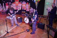 Dzień otwarty w szkole: występ zespołu rockowego na sali gimnastycznej. Uczniowie grają na gitarach elektrycznych, basowych i perkusji. Obok nich stoi grupa wokalna, a w tle widać kolorowe balony, dekoracje z zimowym miasteczkiem oraz drabinki gimnastyczne.