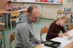 Dzień otwarty w szkole: ojciec towarzyszy córce podczas zajęć w klasie. Dziewczynka skupiona koloruje rysunek pastelami przy szkolnej ławce. W tle widać inne dziecko układające klocki oraz zabawki na parapecie.