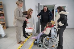 Dzień otwarty w szkole: nauczycielki witają gości przy wejściu. Jedna z nich trzyma wiklinowy koszyk ze słodyczami i częstuje dziewczynkę na wózku inwalidzkim, której towarzyszą rodzice. W tle widać regał z książkami oraz drzwi wejściowe do budynku.