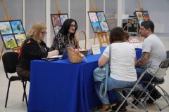 Dzień otwarty w szkole: dwie wicedyrektor szkoły rozmawiają z rodzicami przy stoliku informacyjnym przykrytym niebieskim obrusem. Na stole stoją koszyki ze słodyczami i tabliczka informacyjna, a w tle widać sztalugi z pracami plastycznymi uczniów.