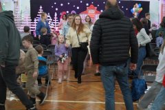 Dzień otwarty w szkole: rodzice i dzieci przemieszczają się po sali gimnastycznej po zakończonej części oficjalnej. Na pierwszym planie widać uśmiechniętą mamę prowadzącą za rękę córkę z pluszakiem. W tle widoczna jest scena z dekoracjami zimowego miasteczka, rzędy krzeseł oraz inni goście wydarzenia.