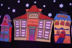 Kolorowa dekoracja sceniczna wykonana z papieru, przedstawiająca bajkowe miasteczko. W centrum znajduje się budynek z napisem „SZKOŁA”, a obok niego fantazyjne kamieniczki w jaskrawych kolorach. Całość umieszczona jest na czarnym tle ozdobionym białymi wycinankami w kształcie płatków śniegu.