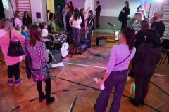 Dzień otwarty w szkole: goście zgromadzeni wokół instrumentów muzycznych na sali gimnastycznej. Na pierwszym planie mała dziewczynka próbuje swoich sił, siedząc przy perkusji, a rodzice robią zdjęcia telefonami. W tle widać młodzieżowy zespół rockowy przygotowujący się do gry oraz innych uczestników wydarzenia.