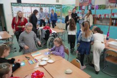 Dzień otwarty w szkole: sala lekcyjna wypełniona dziećmi i rodzicami. Dzieci siedzą przy ławkach, wykonując prace plastyczne, podczas gdy dorośli rozmawiają w głębi sali. Na ścianach widać gazetki szkolne oraz pomoce dydaktyczne.