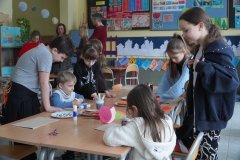 Dzień otwarty w szkole: sala lekcyjna wypełniona dziećmi i rodzicami. Dzieci siedzą przy ławkach, wykonując prace plastyczne, podczas gdy dorośli rozmawiają w głębi sali. Na ścianach widać gazetki szkolne oraz pomoce dydaktyczne.