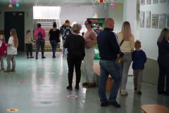 Dzień otwarty w szkole: goście zwiedzają korytarz szkolny. Rodzice i dzieci spacerują wzdłuż ścian, na których wiszą oprawione prace plastyczne uczniów. Na podłodze naklejone są kolorowe litery, a pod sufitem wiszą dekoracyjne lampiony i proporczyki.