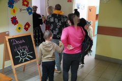 Dzień otwarty w szkole: goście zwiedzają korytarz szkolny. Rodzice i dzieci spacerują wzdłuż ścian, na których wiszą oprawione prace plastyczne uczniów. Na podłodze naklejone są kolorowe litery, a pod sufitem wiszą dekoracyjne lampiony i proporczyki.