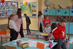 Dzień otwarty w jasnej sali lekcyjnej. Dzieci i dorośli uczestniczą w zajęciach przy stolikach z przyborami plastycznymi oraz na kolorowej macie na podłodze. W tle widać mapę Polski, globus i błękitne szafki z pomocami dydaktycznymi.