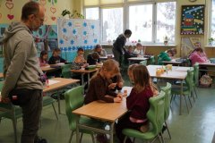 Dzień otwarty w jasnej sali lekcyjnej. Dzieci i dorośli uczestniczą w zajęciach przy stolikach z przyborami plastycznymi oraz na kolorowej macie na podłodze. W tle widać błękitne szafki z pomocami dydaktycznymi.