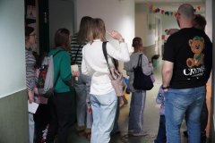 Dzień otwarty w szkole: goście zwiedzają korytarz szkolny. Rodzice i dzieci spacerują wzdłuż ścian, na których wiszą oprawione prace plastyczne uczniów. Na podłodze naklejone są kolorowe litery, a pod sufitem wiszą dekoracyjne lampiony i proporczyki.