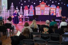 Dzień otwarty w szkole: dyrektor szkoły przemawia do rodziców i dzieci siedzących na widowni w sali gimnastycznej. W tle znajduje się scena z kolorową dekoracją przedstawiającą zimowe miasteczko i choinki z desek.