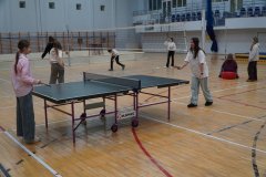 Uczniowie spędzają czas na sportowo w hali gimnastycznej. Na pierwszym planie dwie dziewczynki grają w tenisa stołowego, a w tle widać rozstawioną siatkę do siatkówki oraz inne dzieci ćwiczące na parkiecie.