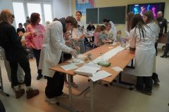 Uczniowie w białych fartuchach przeprowadzają eksperymenty chemiczne w sali lekcyjnej. Na pierwszym planie widać stół z miskami i kartką z napisem „ciecz nienewtonowska”, przy którym dzieci wspólnie badają substancję.
