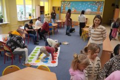 Dzieci i rodzice w sali świetlicowej podczas dnia otwartego. Na pierwszym planie dwoje dzieci gra w Twistera na podłodze, a w głębi pozostali goście siedzą przy stolikach lub oglądają dekoracje ścienne o tematyce zimowej.