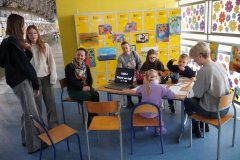 Grupa dzieci i dorosłych siedzi przy stoliku z kredkami i laptopem na tle żółtych szafek szkolnych ozdobionych pracami plastycznymi. Na ekranie widnieje napis „Bajka Vincent van Gogh”, a uczestnicy wspólnie biorą udział w warsztatach kreatywnych.