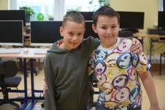 Dwaj uśmiechnięci chłopcy pozują do zdjęcia, obejmując się ramionami w sali komputerowej. W tle widać rzędy biurek z monitorami i klawiaturami.