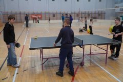 Uczniowie spędzają czas na sportowo w hali gimnastycznej. Na pierwszym planie dzieci grają w tenisa stołowego, a w tle widać rozstawioną siatkę do siatkówki oraz inne dzieci ćwiczące na parkiecie.