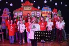 Występ chóru szkolnego podczas dnia otwartego. Grupa uczniów w czerwonych i białych koszulkach z logo „Chór Tutu” śpiewa na scenie. Na pierwszym planie dziewczyna śpiewa do mikrofonu, a w tle widać kolorowe dekoracje przedstawiające budynki.