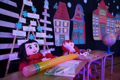 Dekoracja teatralna na szkolnej scenie przedstawiająca dwie duże kukły siedzące w ławkach. Przed nimi leżą podręczniki i ogromny żółty ołówek, a w tle widać kolorowe kartonowe kamienice, globus oraz choinkę z wyrazami kojarzącymi się z zimą.