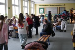 Główny hol Szkoły Podstawowej nr 22 wypełniony gośćmi podczas dnia otwartego. Rodzice i dzieci siedzą na ławkach lub stoją przy stolikach, a na żółtej ścianie w tle widnieje nazwa placówki oraz cytat Janusza Korczaka.